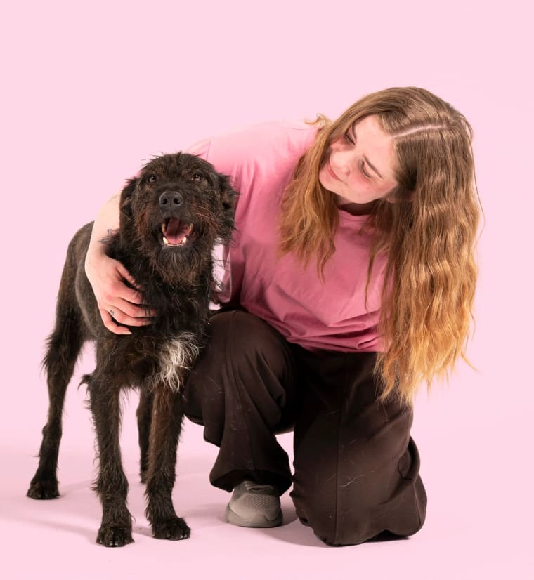 Ulrika Winge sitter på knä och kramar en svart lockig hund mot en rosa bakgrund