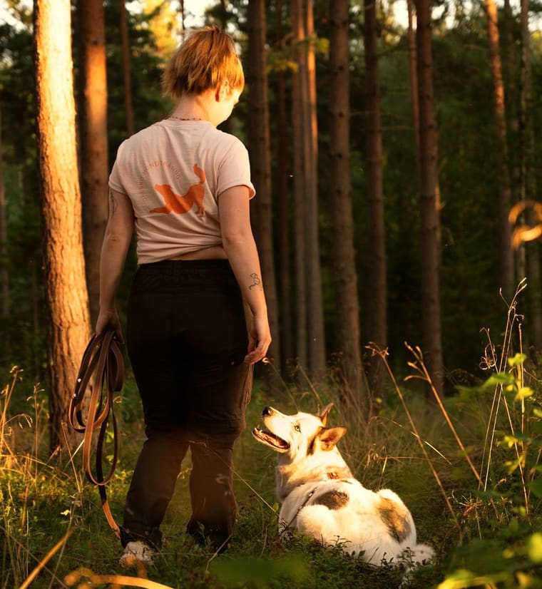 Ulrika Winge går i en solbelyst skog och tittar ner på en vit och brun hund som ligger i gräset