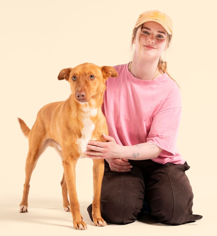 Ulrika Winge i keps sitter på knä bredvid en ljusbrun hund och ler mot kameran