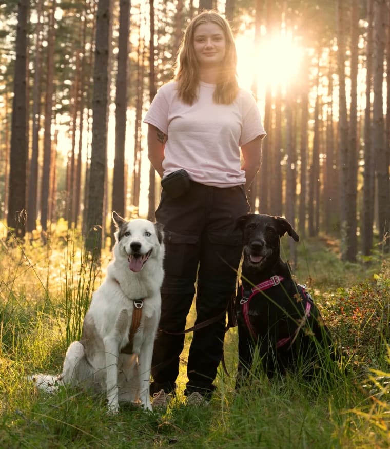 Ulrika Winge står i en solbelyst tallskog mellan en vit och en svart hund